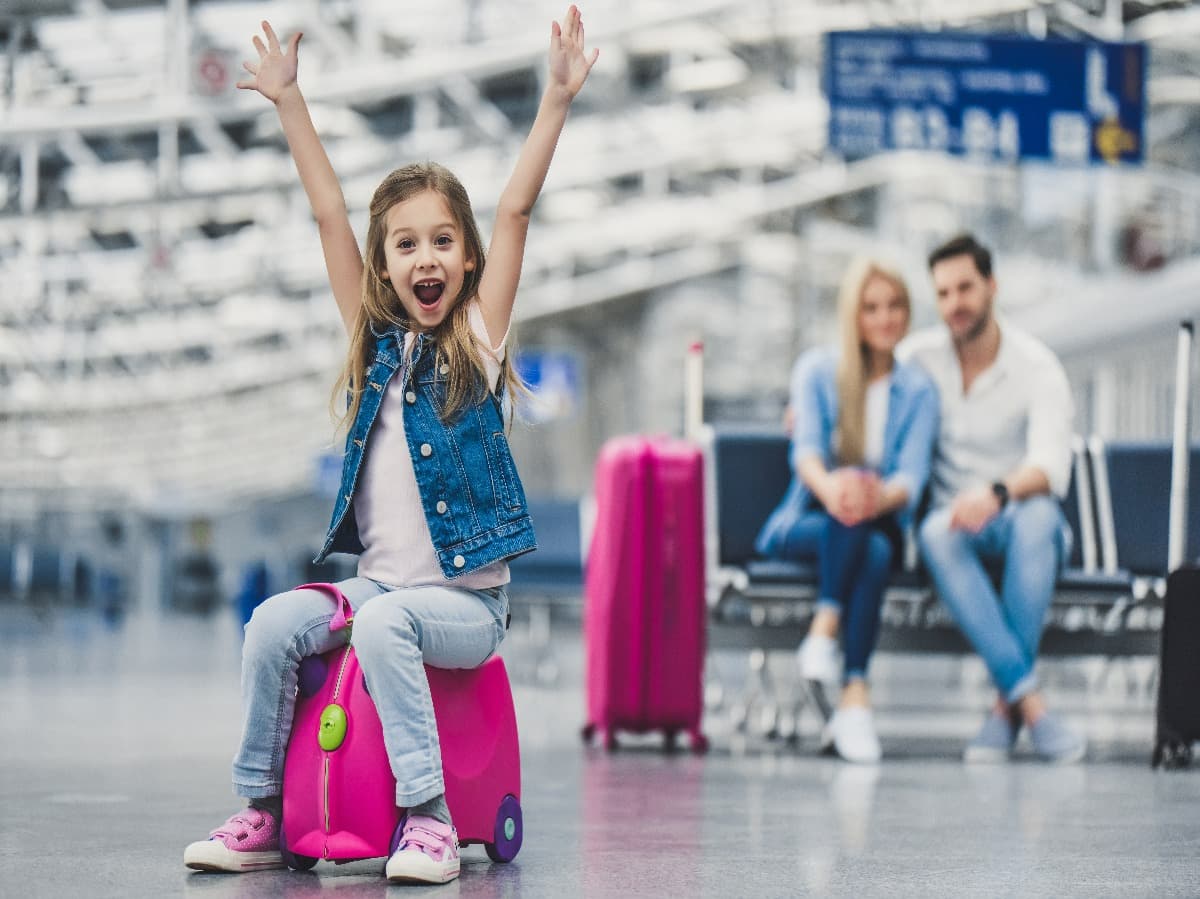 chica sentada en una maleta en el aeropuerto con sus padres mirando al fondo